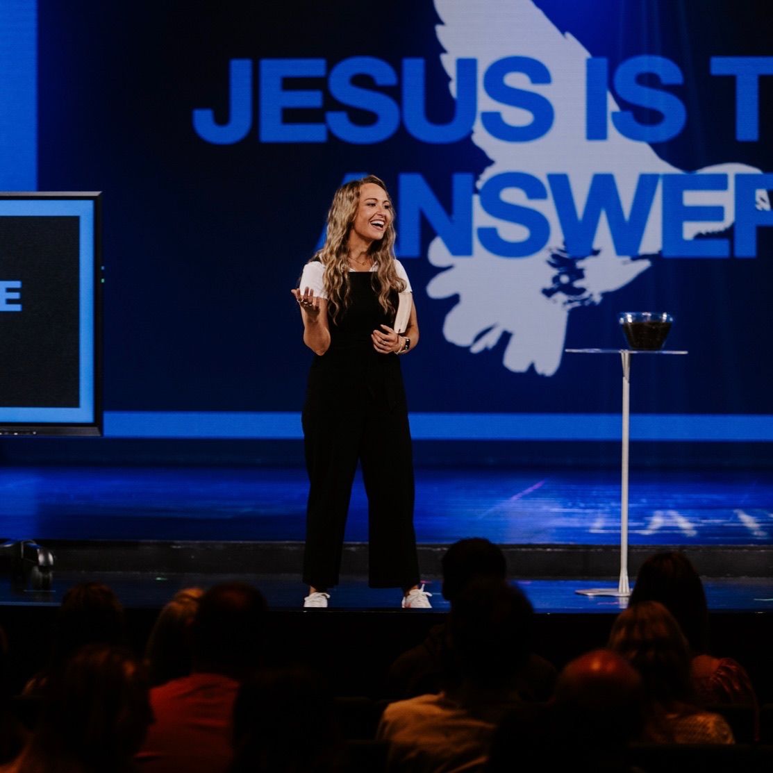 Kate Downing speaking on stage in front of a screen reading “Jesus is the answer”